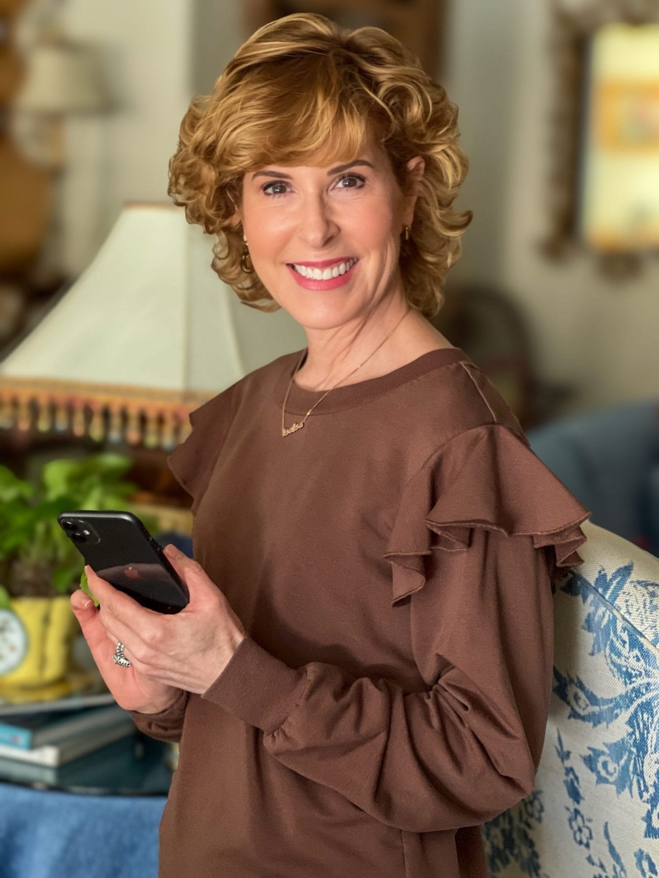 woman standing in living room wearing brown ruffle shoulder sweatshirt holding a smartphone