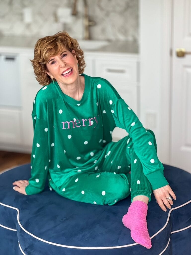 woman in green and white polka dot velour pajamas with word Merry on shirt and pink socks sitting on an ottoman