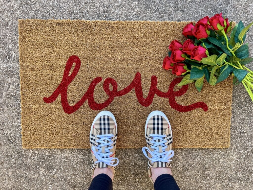 woman's feet in burberry sneakers standing on love doormat with red silk roses to the side