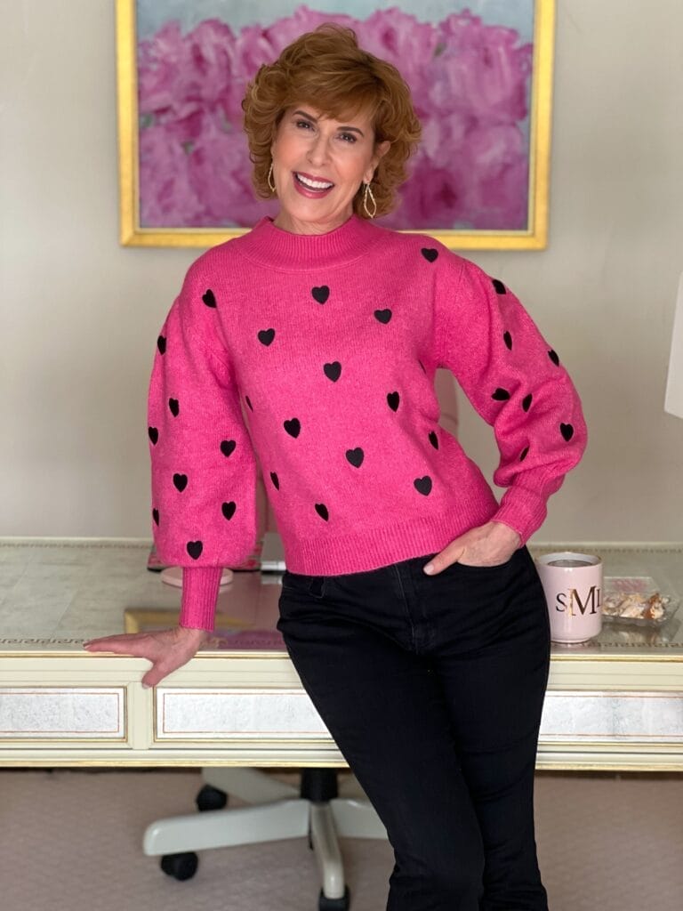 woman wearing pink and black hearts sweater from shop avara with one hand in pocket leaning against desk
