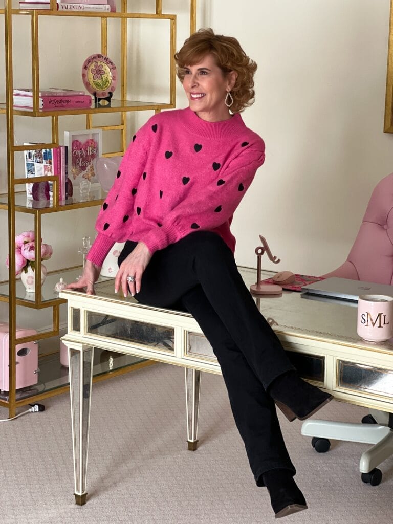 woman sitting on a desk in her office wearing a pink sweater with black hearts and black jeans and booties.
