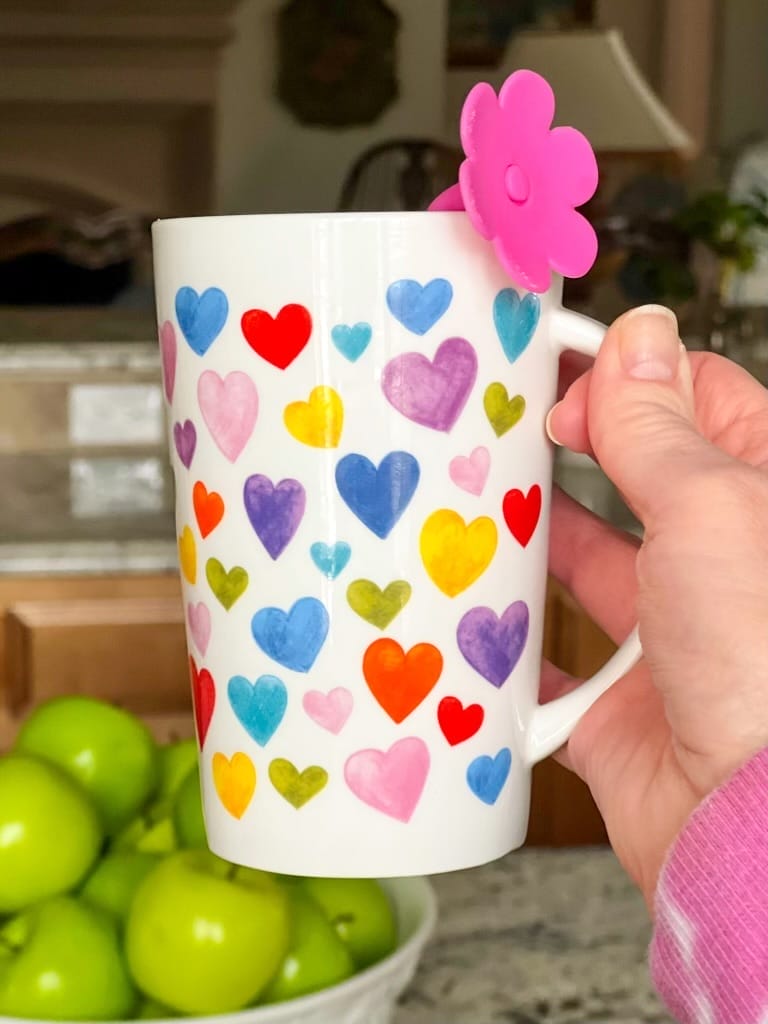 Woman's hand holding a heart mug with a pink flower tea infuser in it and a bowl of apples in the background