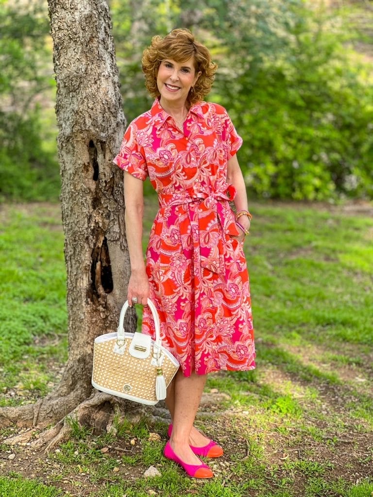 Woman standing next to a tree in pink and orange shirt dress holding a cane and white handbag