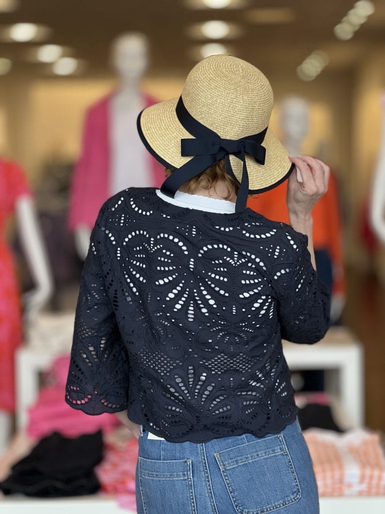 woman dressed in white tee and crochet navy 3/4 sleeve jacket wearing straw hat from back