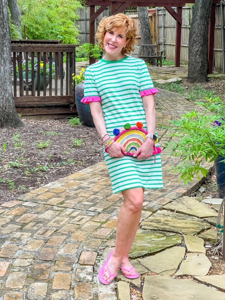 Woman standing outside in green and white striped dress with hot pink ruffle on sleeves holding colorful rattan bag and wearing pink flip flops