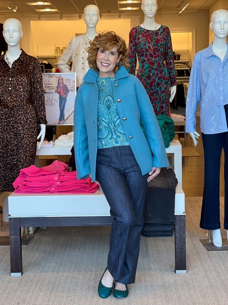 woman over 50 standing in a talbots store wearing blue coat over cashmere blue print sweater and trouser jeans with teal ballet flats