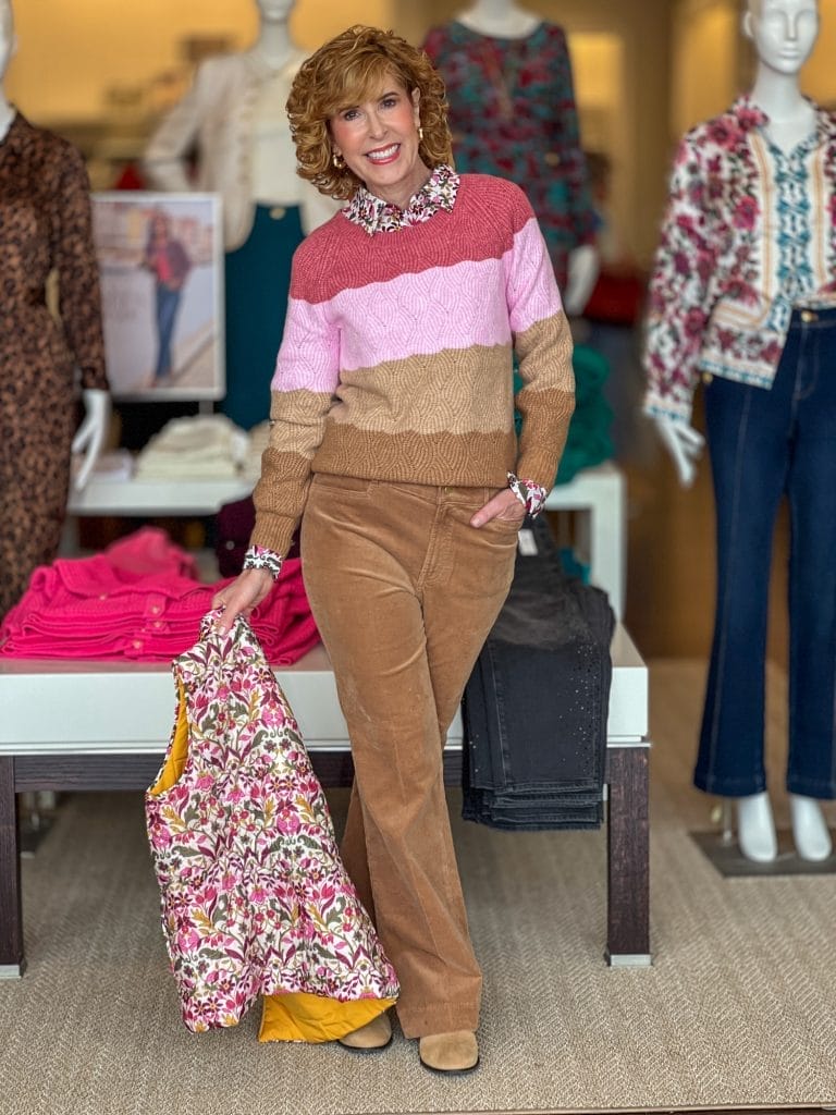 woman over 50 standing in a talbots store wearing striped sweater with brown corduroy pants and holding a floral vest