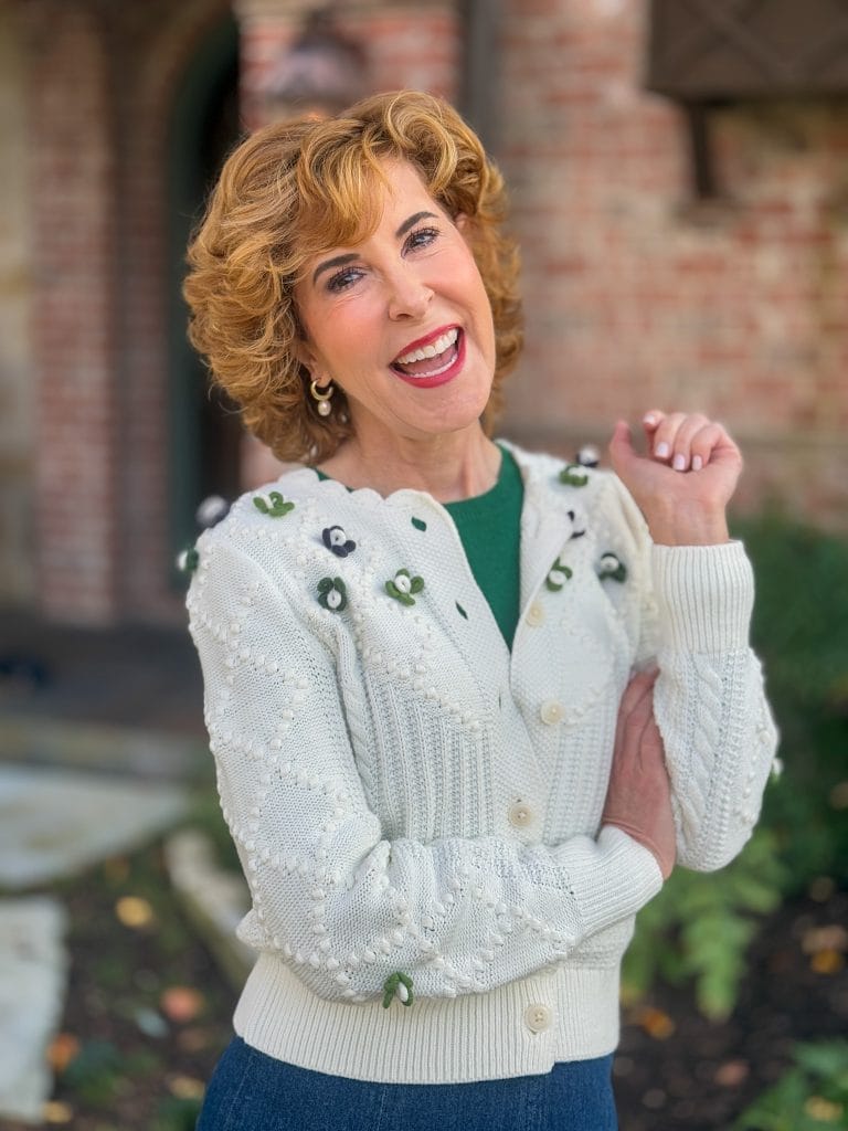 woman over 50 wearing off white cardigan with blue and green 3d flowers standing in front of a house